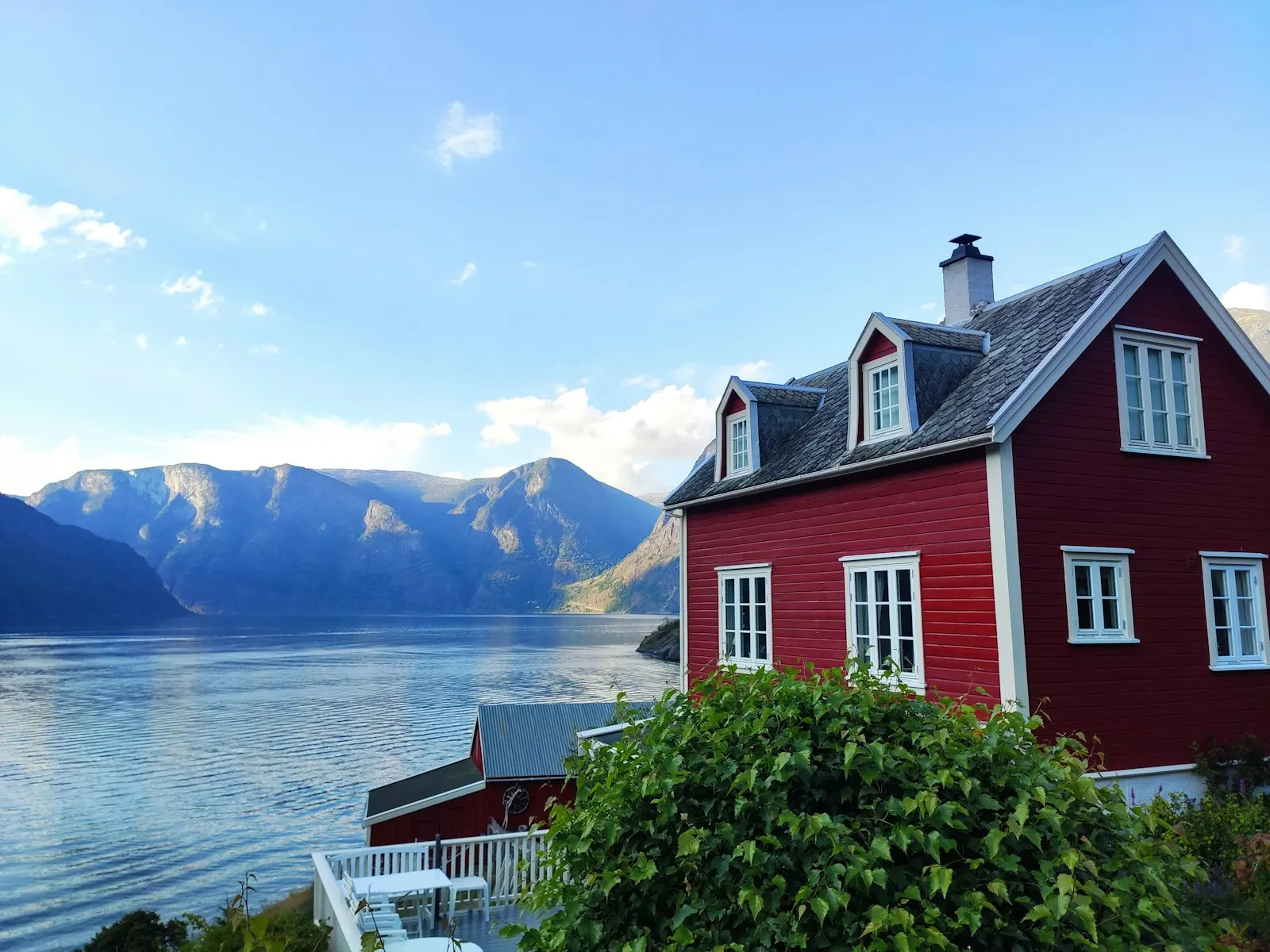 Rødt trehus med hvite vinduer i en hage rett over Aurlandsfjorden, med blå fjell og sommerhimmel
